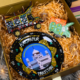 A gift box filled with shredded paper contains a round black decorative plate featuring a blue and white illustration of a mosque with 'Kartarpur Pakistan' text, plus two colorful painted truck-shaped ornaments.