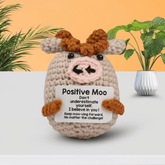 Crocheted cow toy holding a motivational card with plants in the background