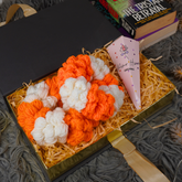 A gift box containing handmade orange and white yarn pom-poms, a pink cone-shaped gift bag with a butterfly design, and a black book on a gray textured rug.