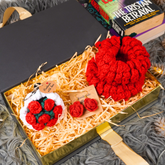 A gift box filled with handmade crochet items, including a large red crocheted flower, a white and red flower ornament, a pair of red crochet earrings, and cards, set on a gray shag rug with books in the background.