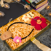 A gift box filled with shredded paper contains a black headband with pink and cream crochet flowers, a red crochet flower keychain, and two cream crochet hair clips. The box is styled on a gray textured blanket with books and fairy lights in the background.