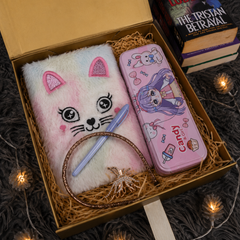 A curated gift box set containing a fluffy pastel pink and white cat-face notebook, a pink cartoon pencil case, a glittery gold ring, and a light blue pen, arranged in brown paper shred inside an open gold box with decorative books and fairy lights.