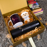 A curated gift box set from Sentimental Creations Co containing a white ceramic mug with a dark purple gradient design, a black insulated tumbler, and a small knitted owl plush toy, all nestled in shredded paper inside a gold box. A book titled 'The Tristan Betrayal' by Robert Ludlum is placed next to the box on a gray textured rug with warm fairy lights.