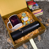 A curated gift box from Sentmental Creations Co. containing a black insulated tumbler, a white ceramic mug with a dark purple printed design, and a small crocheted green character toy, all nestled in shredded paper inside a gold box.