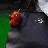 Handmade brown crochet bean keychain with a smiling face hanging on a Coach leather bag.