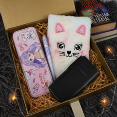A curated gift box set containing a pink cartoon anime pencil case, a white plush cat face notebook, a light blue pen, and a black wallet, arranged in brown paper shred inside an open cardboard box.