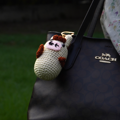 Adorable beige and brown handmade crochet cow plush keychain holding a small motivational sign, shown hanging on a black designer handb