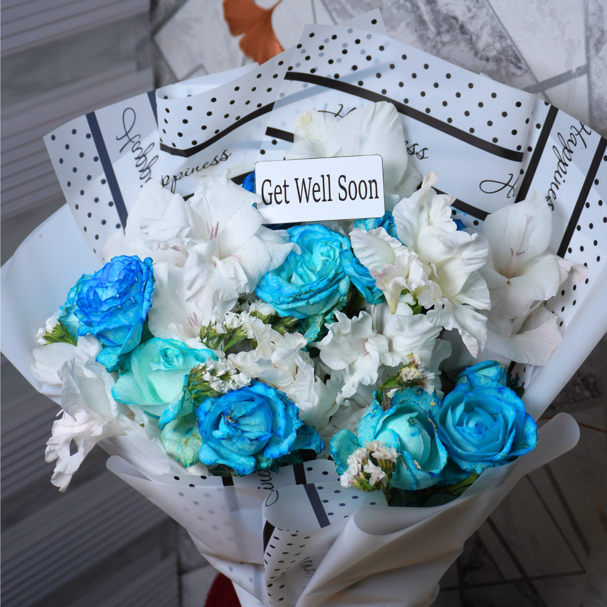 Bouquet of blue and white flowers with a 'Get Well Soon' card on a tiled floor.