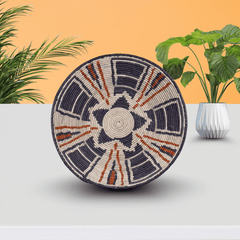 Decorative woven basket with geometric pattern on a light gray surface with plants in the background
