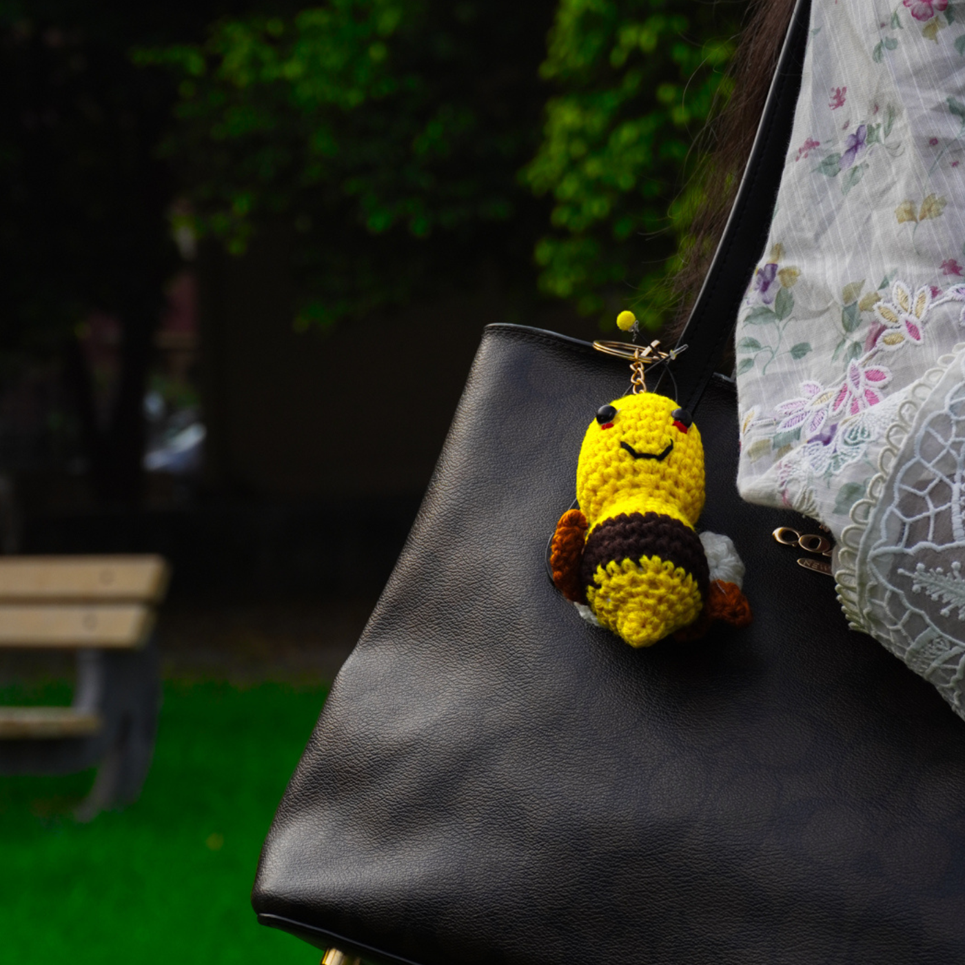 Handmade yellow crochet motivational bee plush keychain hanging on a black handbag outdoors in Lahore, Pakistan.